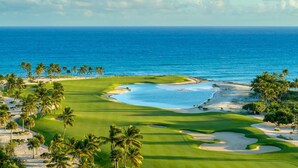 A golf course with a water hazard, palm trees, and a view of the ocean. - Cozy 1br w Balcony Pool Beach Access Cap Cana (Punta Cana)
