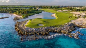 A coastal golf course with a water body, green fairways, and sand bunkers. - Cozy 1br w Balcony Pool Beach Access Cap Cana (Punta Cana)