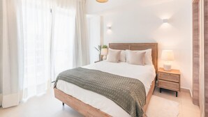 A bedroom with a wooden bed, white bedding, a bedside table with a lamp, and a window with sheer curtains. - Cozy 1br w Balcony Pool Beach Access Cap Cana (Punta Cana)