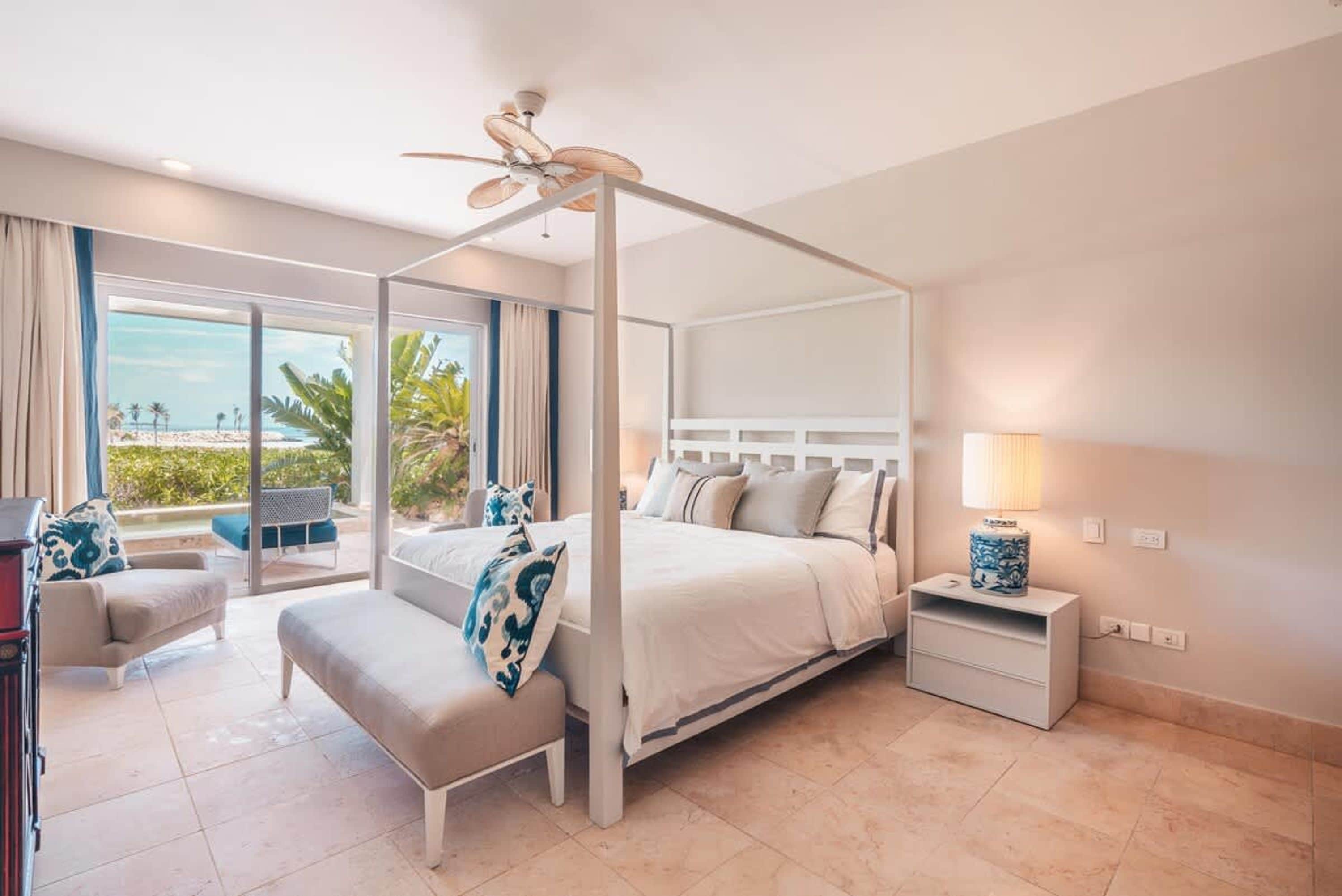 Een bed met hemelbed, een plafondventilator, een nachtkastje met lamp, een bankje en uitzicht op het strand.