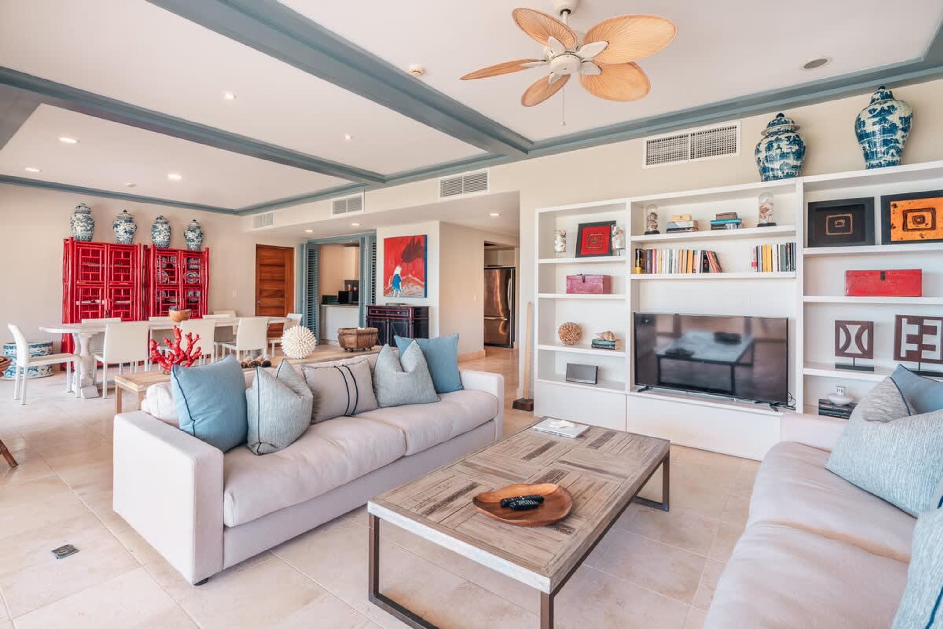 Un salón amplio con un sofá, una mesa de centro y un televisor. Se observan estanterías con objetos decorativos y un comedor al fondo.