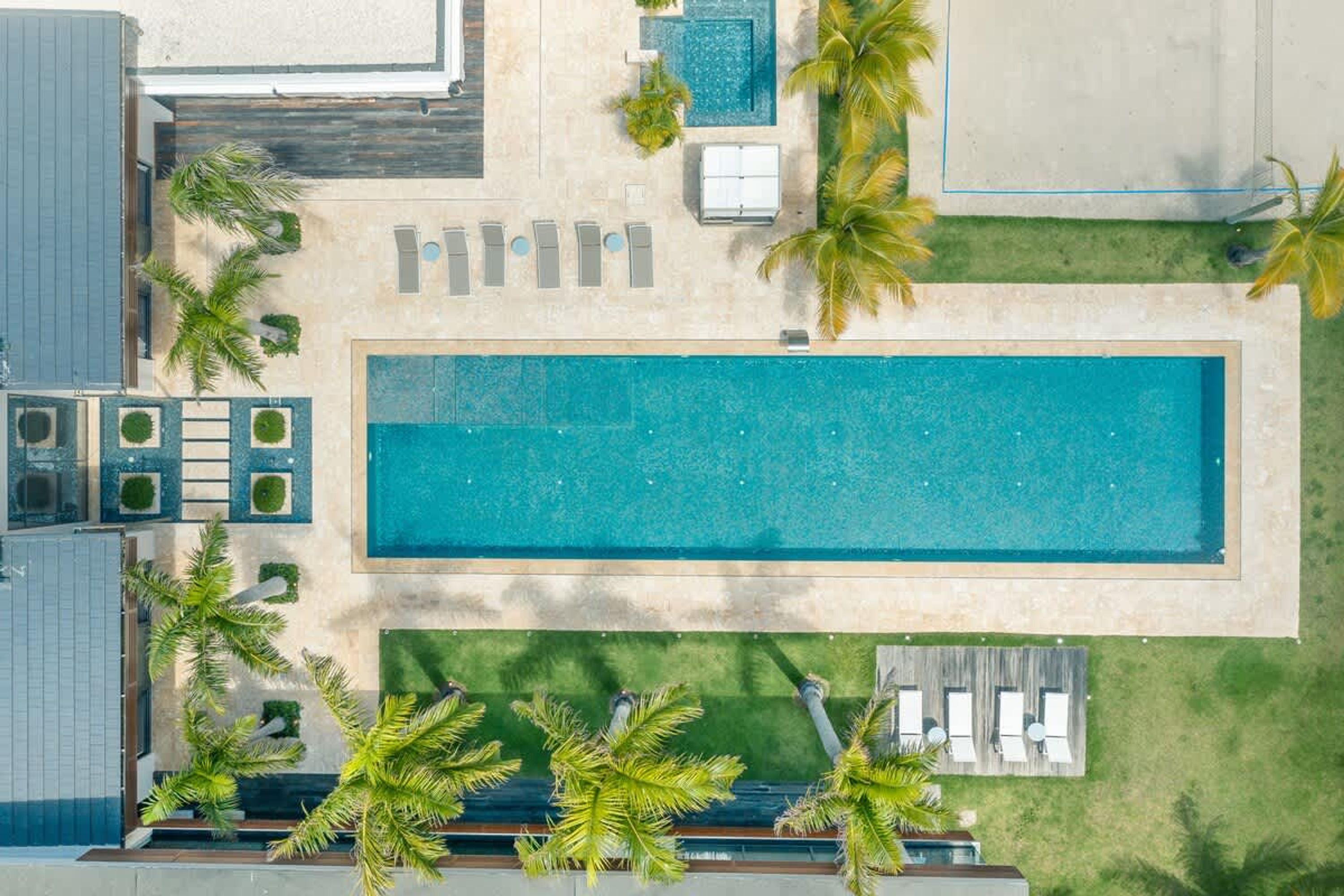 An aerial view of a resort with a rectangular swimming pool, palm trees, and lounge chairs.