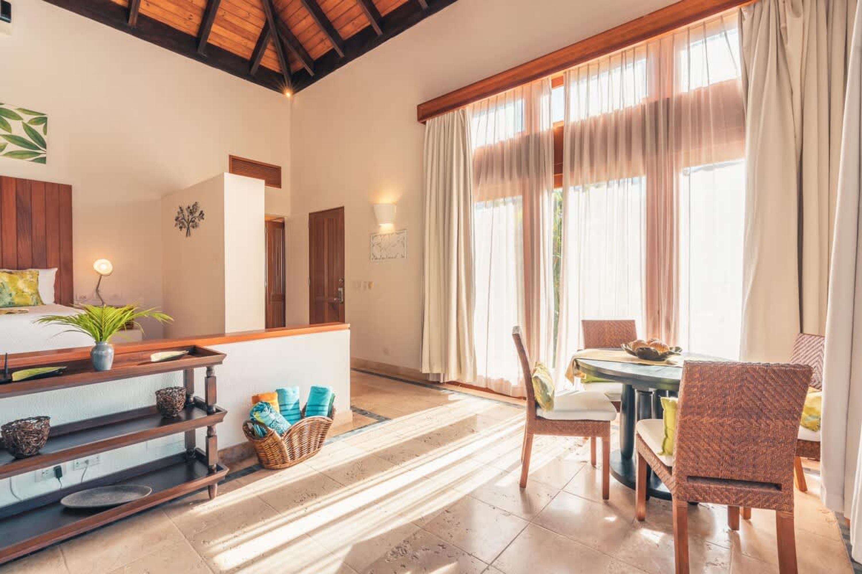 A room with a wooden ceiling, a dining table with chairs, a plant, and a shelf with baskets.