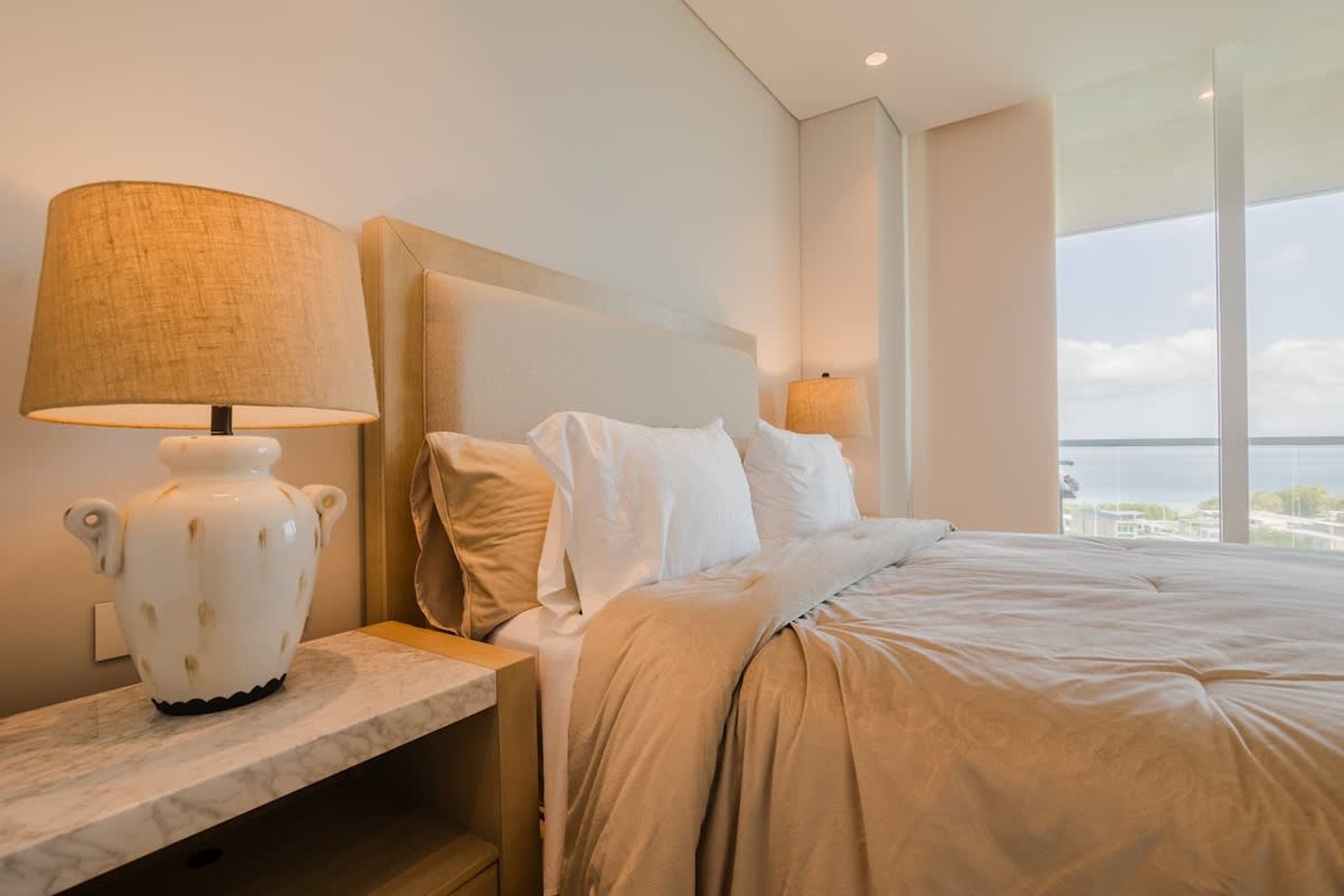 A modern hotel room with a large bed, a nightstand with a lamp, and a view of the sea.