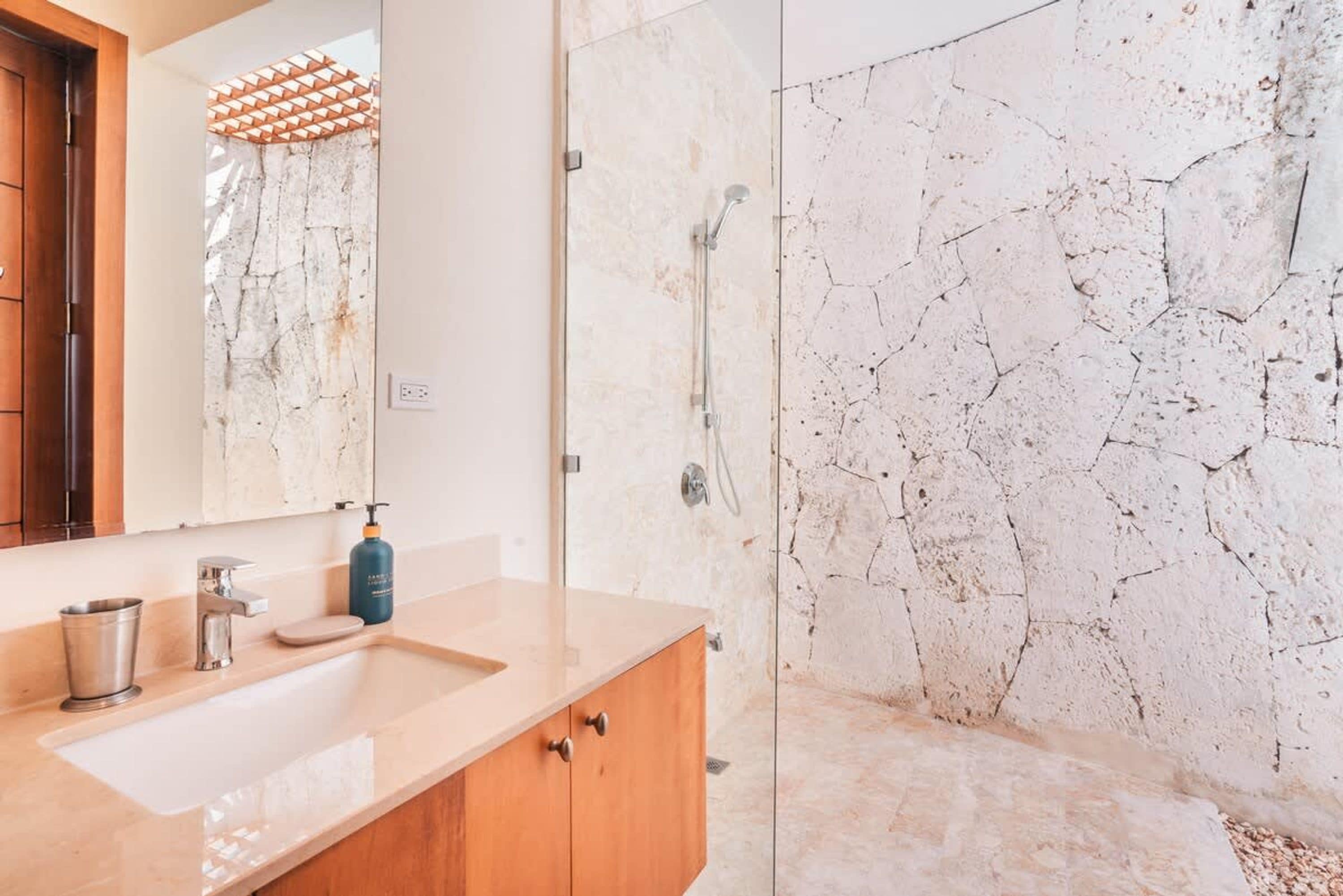 A bathroom with a marble sink, wooden cabinets, a glass shower enclosure, and a stone wall.