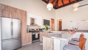A modern kitchen with wooden cabinets, stainless steel appliances, a central island with bar stools, and a high ceiling with exposed beams.