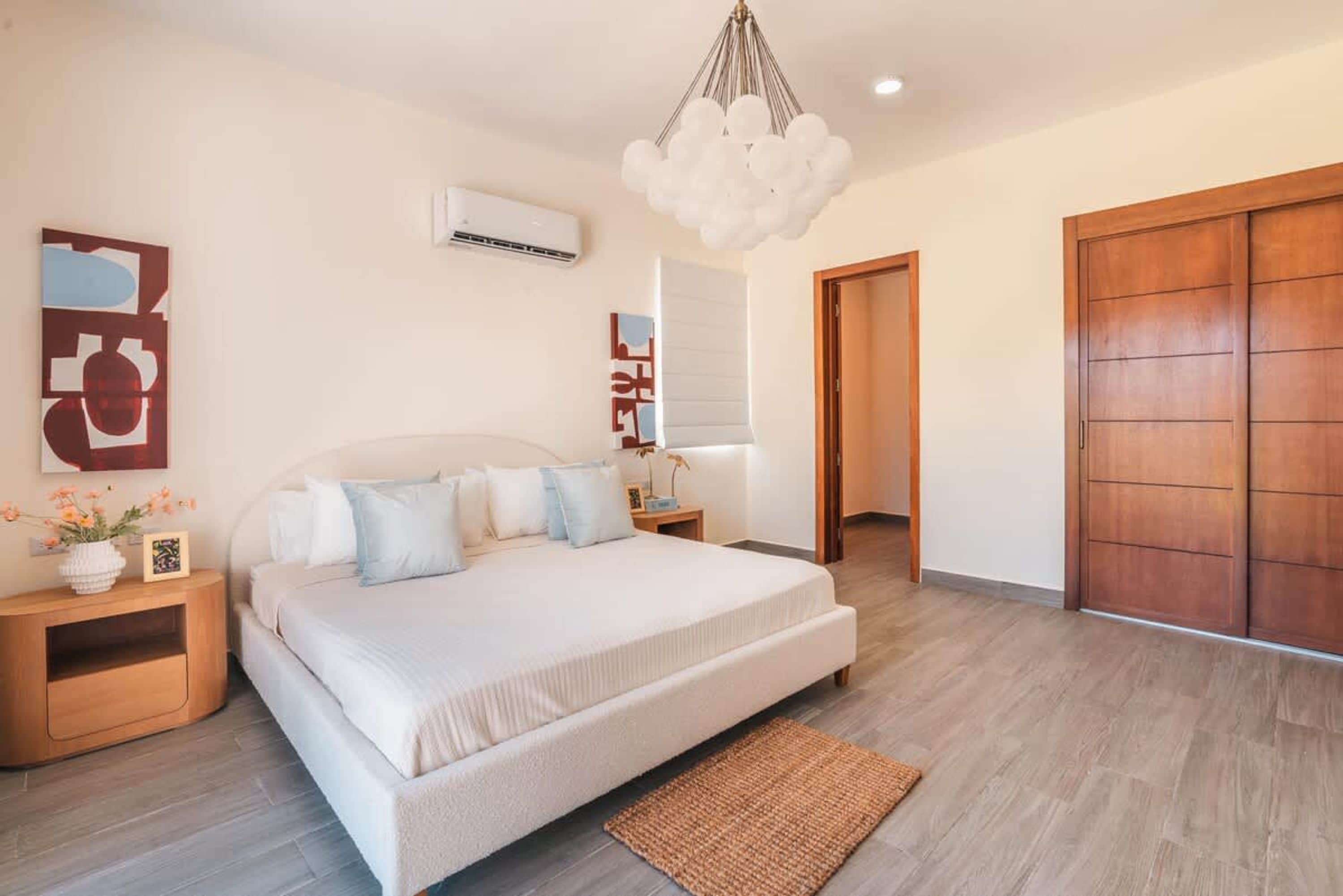 A bedroom with a bed, bedside tables, a hanging light fixture, an air conditioning unit, and a wooden wardrobe.