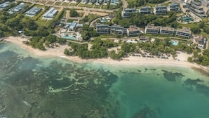 Vista aérea de un complejo turístico costero con varios edificios, una playa y aguas cristalinas.