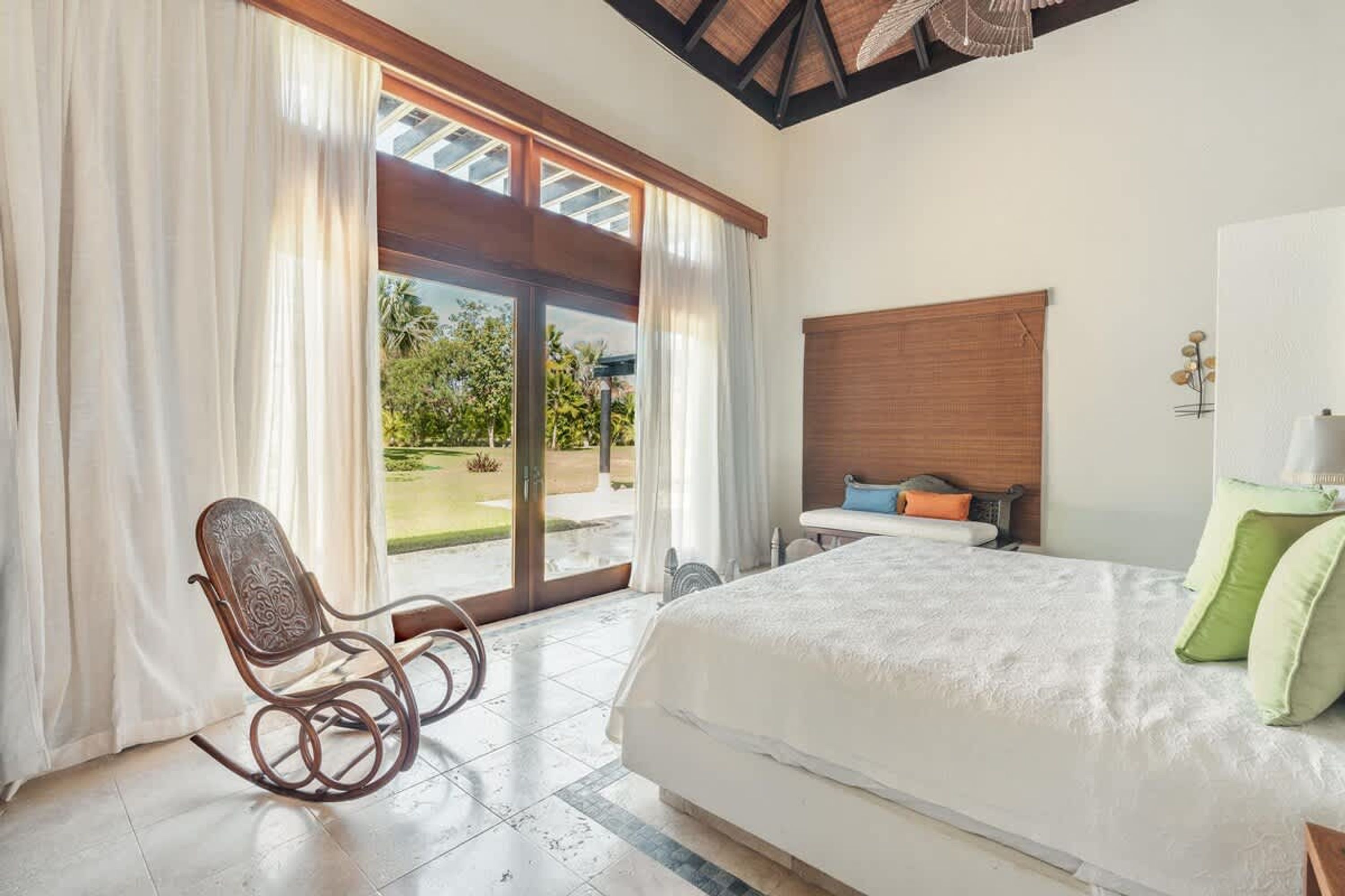 A bedroom with a large bed, a rocking chair, a small table, and a view of the outdoors.