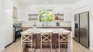 Uma cozinha moderna com ilha central, banquetas, geladeira e janela com vista para o verde.