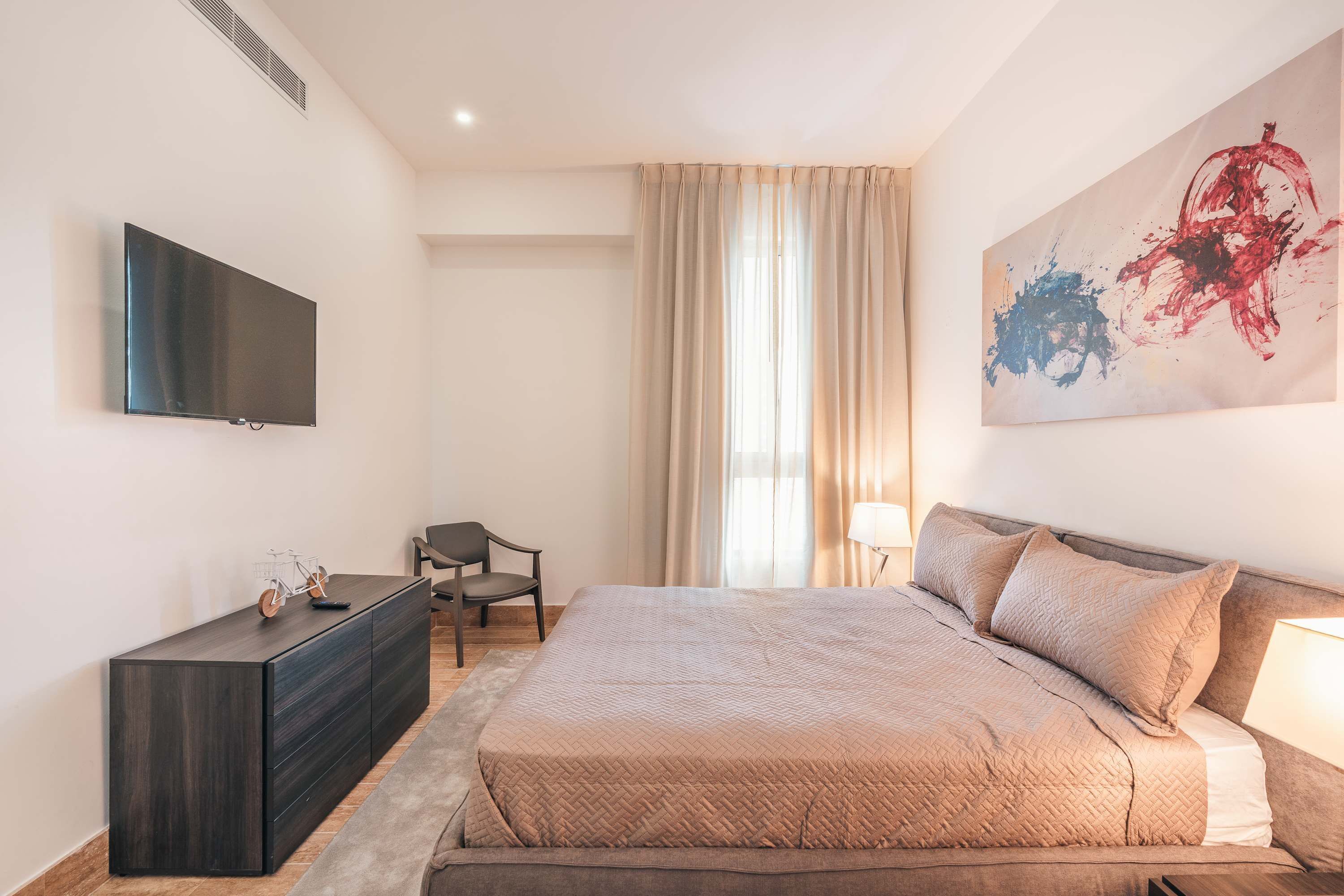 A bedroom with a bed, a TV, a desk, a chair, and a large abstract painting.