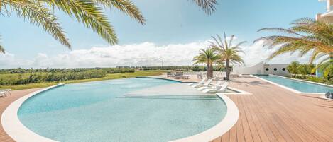 A pool area with a wooden deck, lounge chairs, palm trees, and a view of distant buildings.