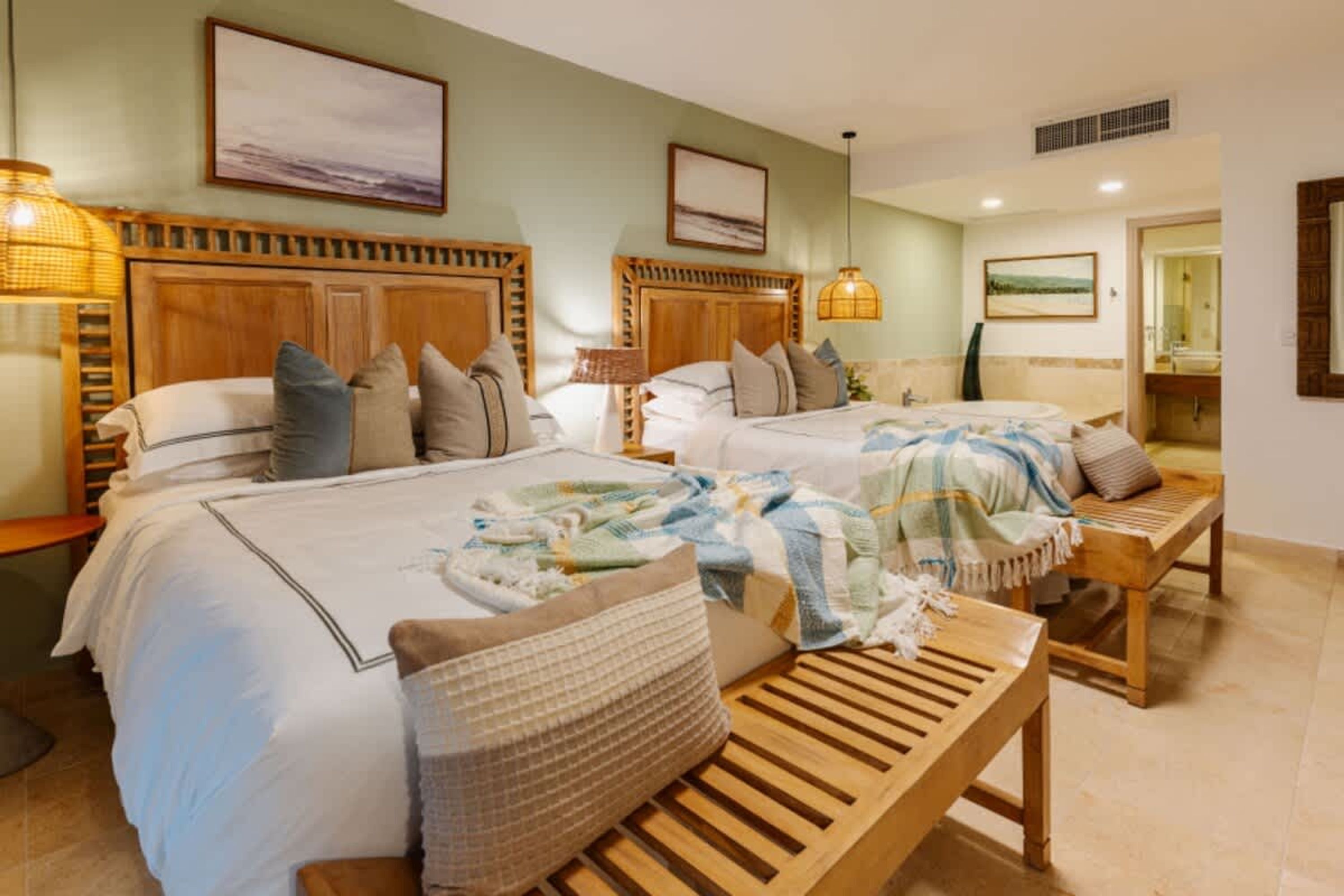 Habitación de hotel con dos camas, un banco de madera y un baño visible a través de una puerta abierta.