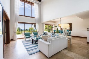 Uma sala de estar espaçosa, com um sofá, cadeiras, uma mesa de centro de vidro e vista para o exterior.