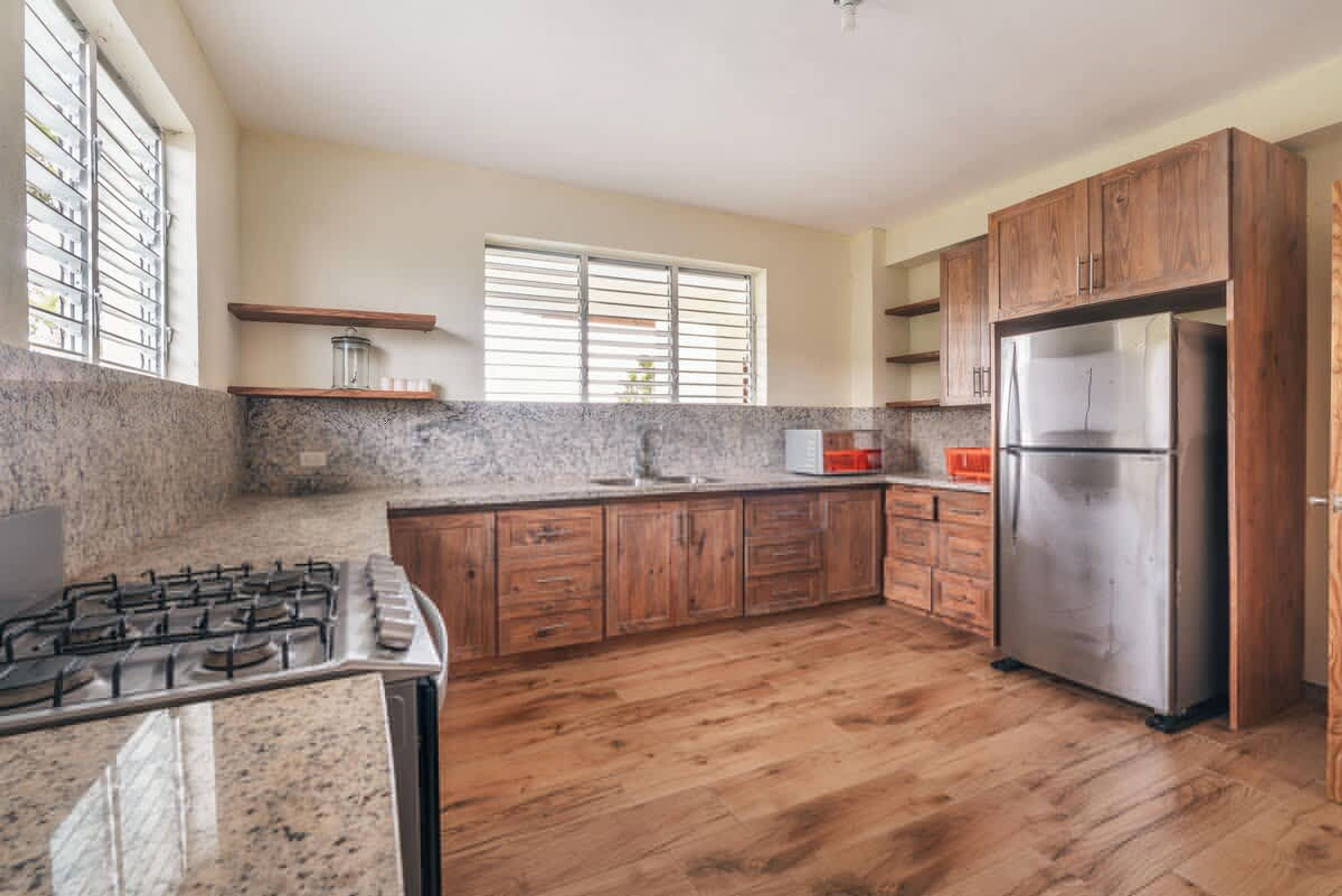 A kitchen with wooden cabinets, a stainless steel refrigerator, a granite countertop, and a window with blinds.