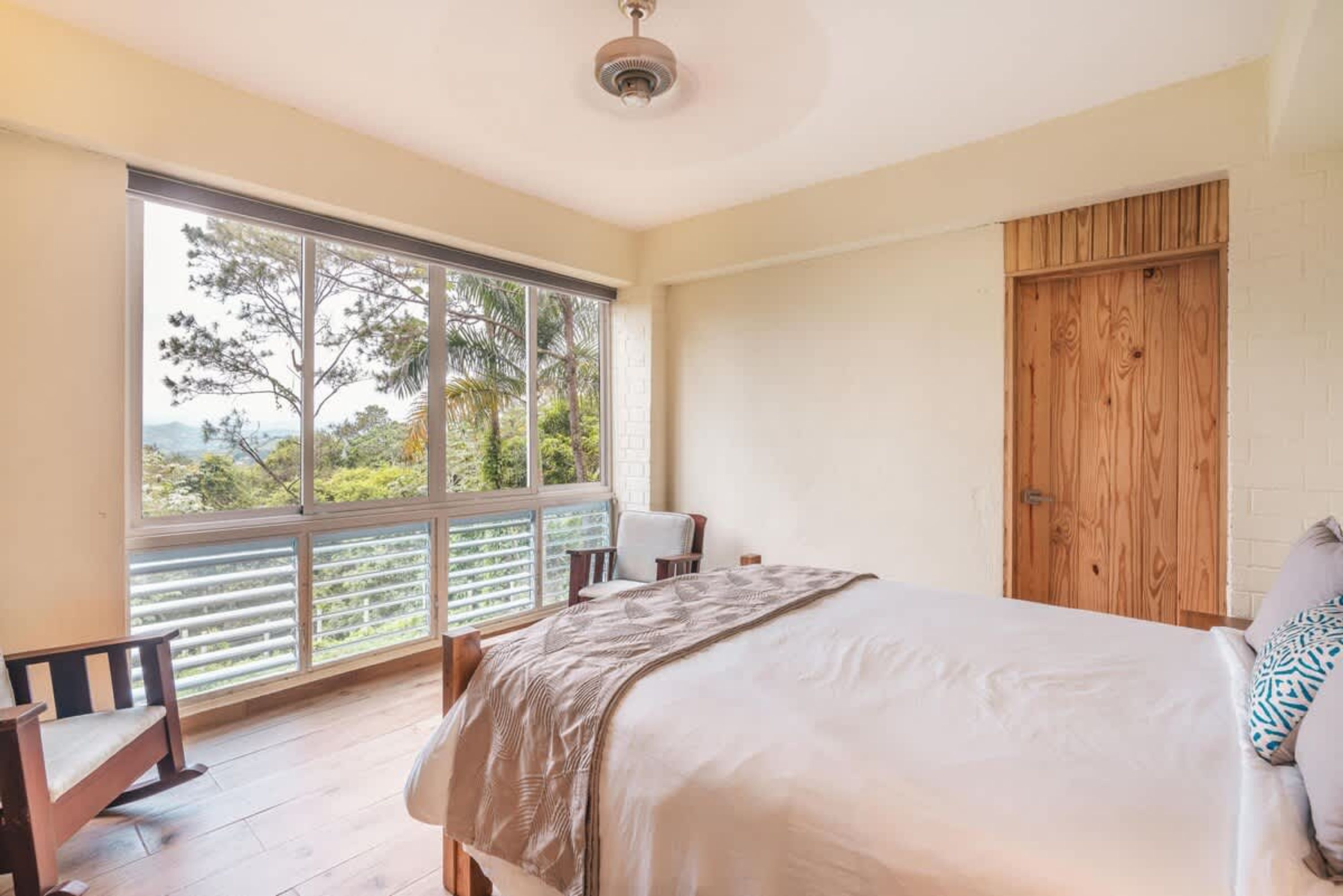 A bedroom with a large window, a bed, a chair, and a wooden wardrobe.