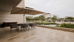 An outdoor patio with a table and chairs, a large umbrella, and a view of a swimming pool and greenery. - Two Bedrooms apt With Views in Baru With Jacuzzi (Cartagena)