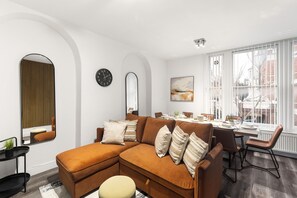 A modern living room with a sectional sofa, dining table, chairs, a large mirror, and a window with a view. - Cozy 2-bedroom Flat in Central London (London)