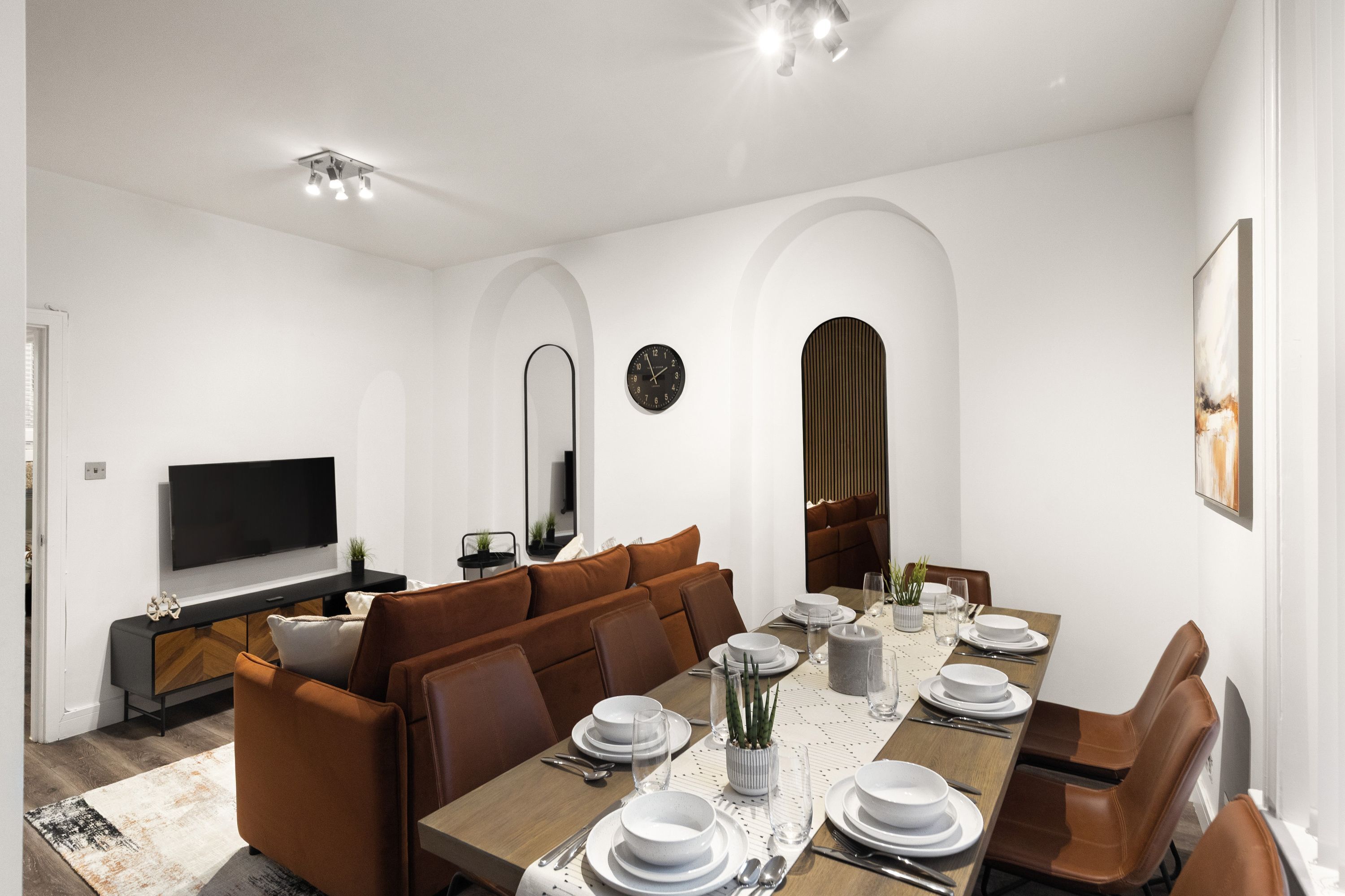 A modern dining room with a long table set for six, a brown sofa, and a flat-screen TV on a black console.