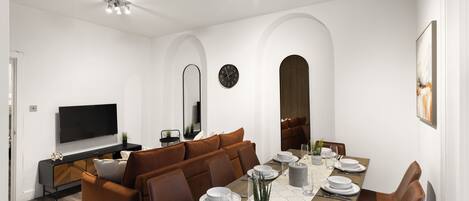 A modern dining room with a long table set for six, a brown sofa, and a flat-screen TV on a black console.