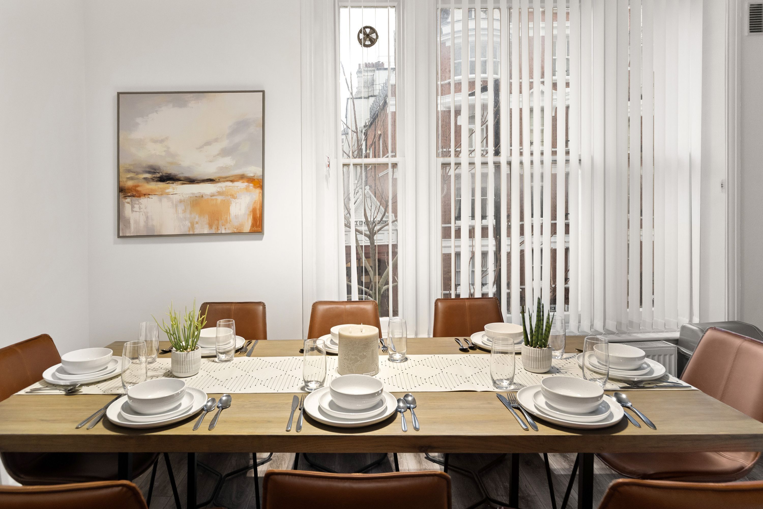 Een eetruimte met een houten tafel voor zes personen, witte borden, glazen en bestek. Aan de muur hangt een schilderij, er is een raam met zonwering en een uitzicht op gebouwen.