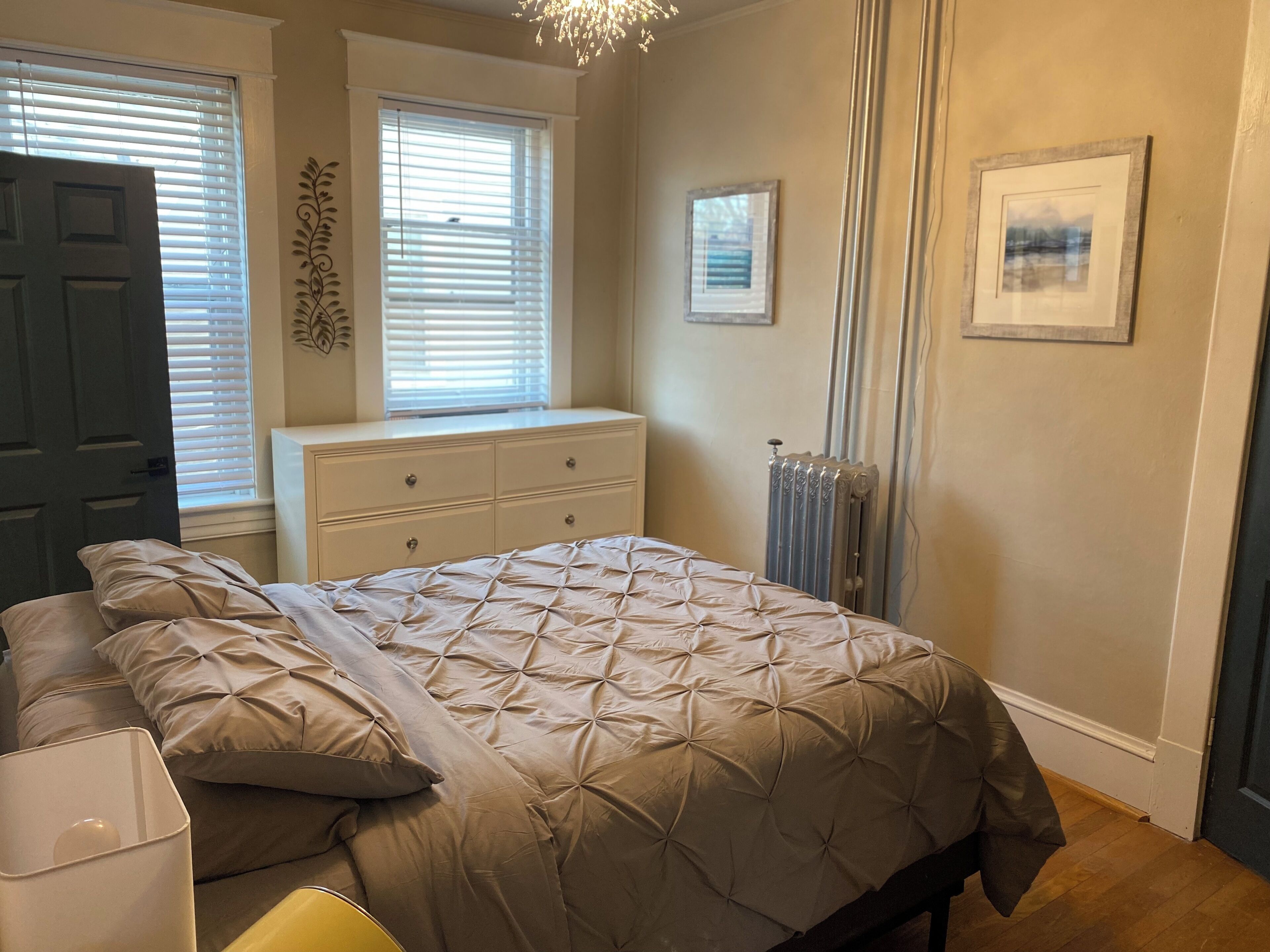 A bedroom with a bed, dresser, radiator, and two windows with blinds.