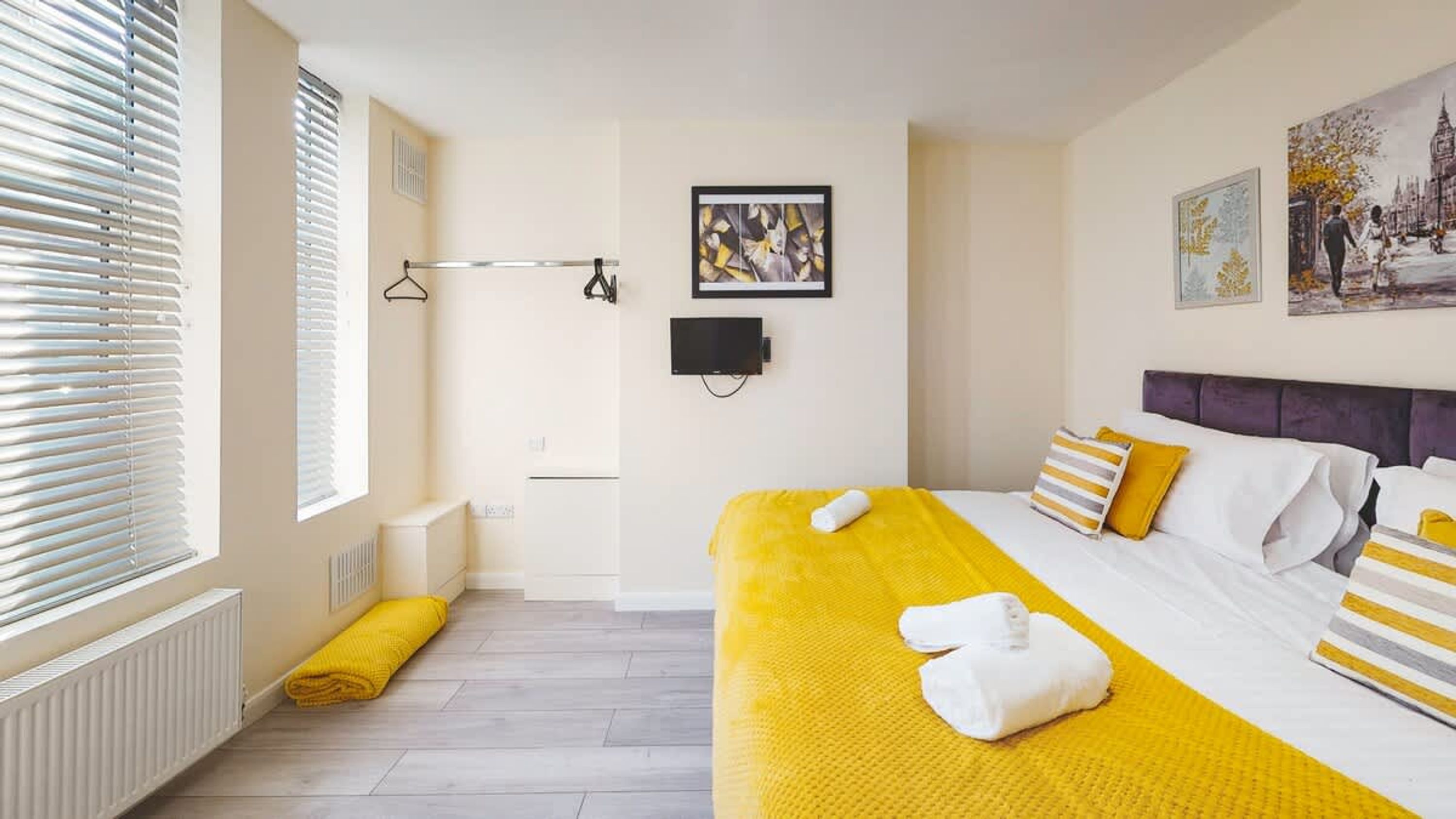 A hotel room with a bed, a window with blinds, a radiator, a towel, and a painting on the wall.