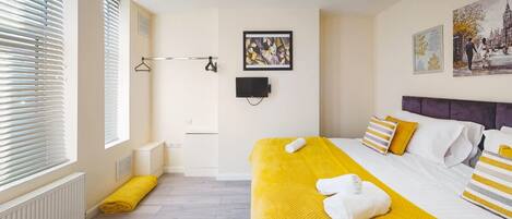 A hotel room with a bed, a window with blinds, a radiator, a towel, and a painting on the wall.