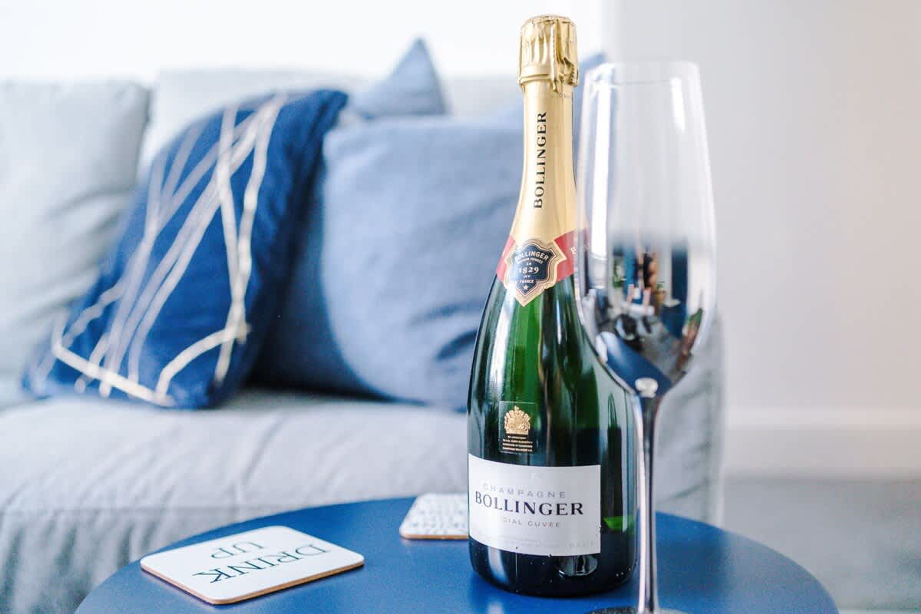 Une bouteille de champagne Bollinger et un verre posés sur une table bleue, dans une pièce où se trouve un coussin aux motifs bleus et blancs.