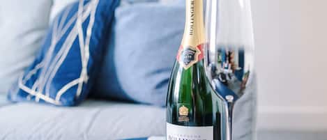 Une bouteille de champagne Bollinger et un verre posés sur une table bleue, dans une pièce où se trouve un coussin aux motifs bleus et blancs.