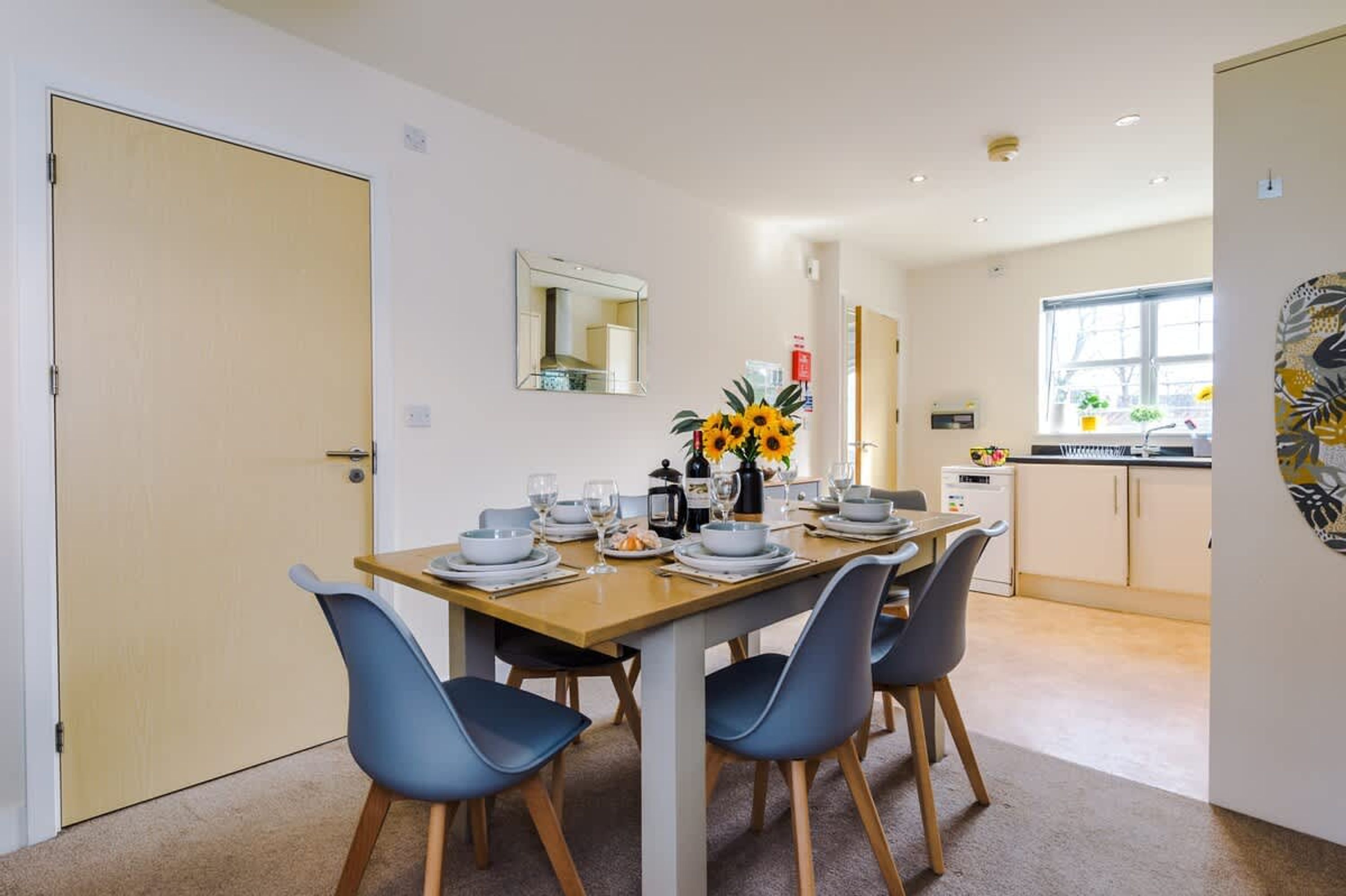 A dining area with a table set for four, a vase of sunflowers, and a kitchen in the background.