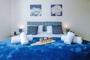Ein ordentlich bezogenes Bett mit einer blauen Decke, zwei Kissen mit geometrischem Muster und einem Holztischchen mit drei Donuts.