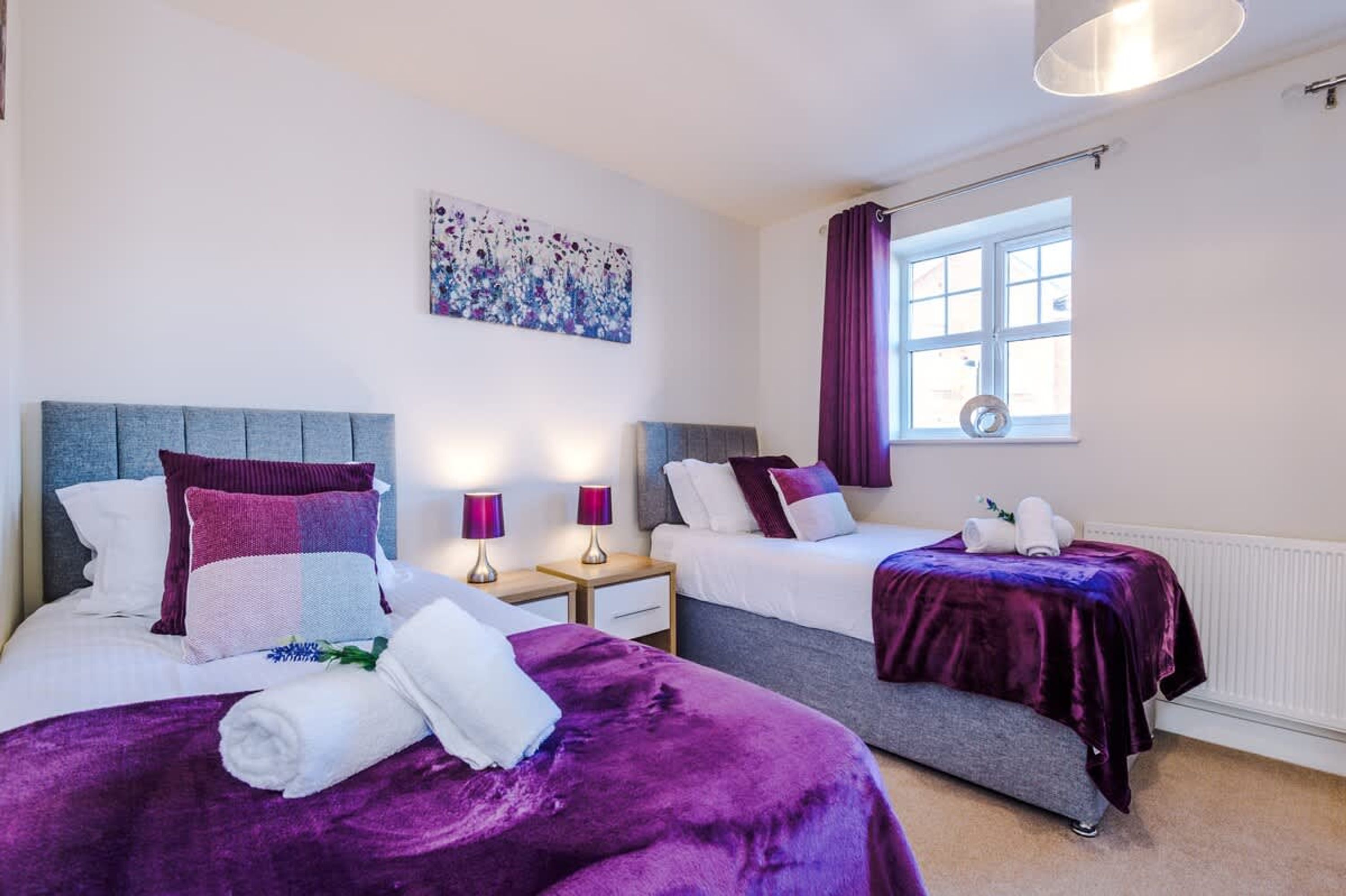A hotel room with two beds, a window with curtains, a wall-mounted artwork, and a bedside table with lamps.