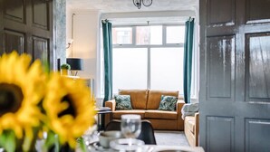 Ein Essbereich mit Tisch für vier Personen, einer Couch im Hintergrund und einem Fenster mit Vorhängen.