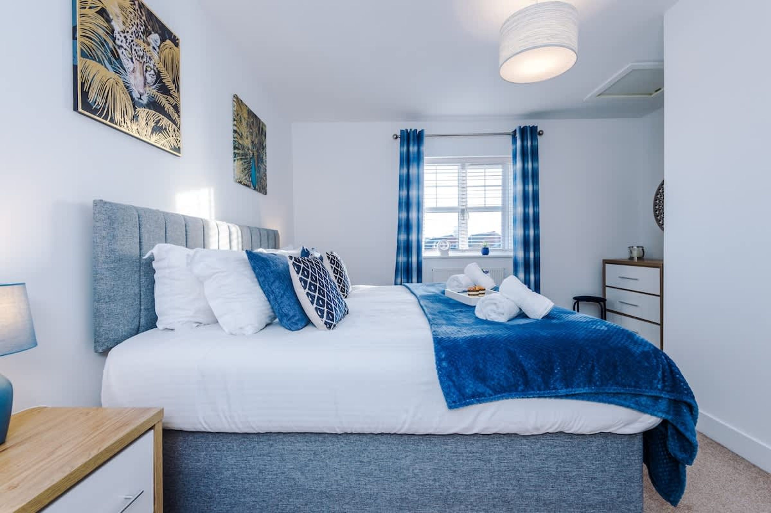 A bedroom with a bed, a nightstand, a dresser, a window with curtains, and wall art.