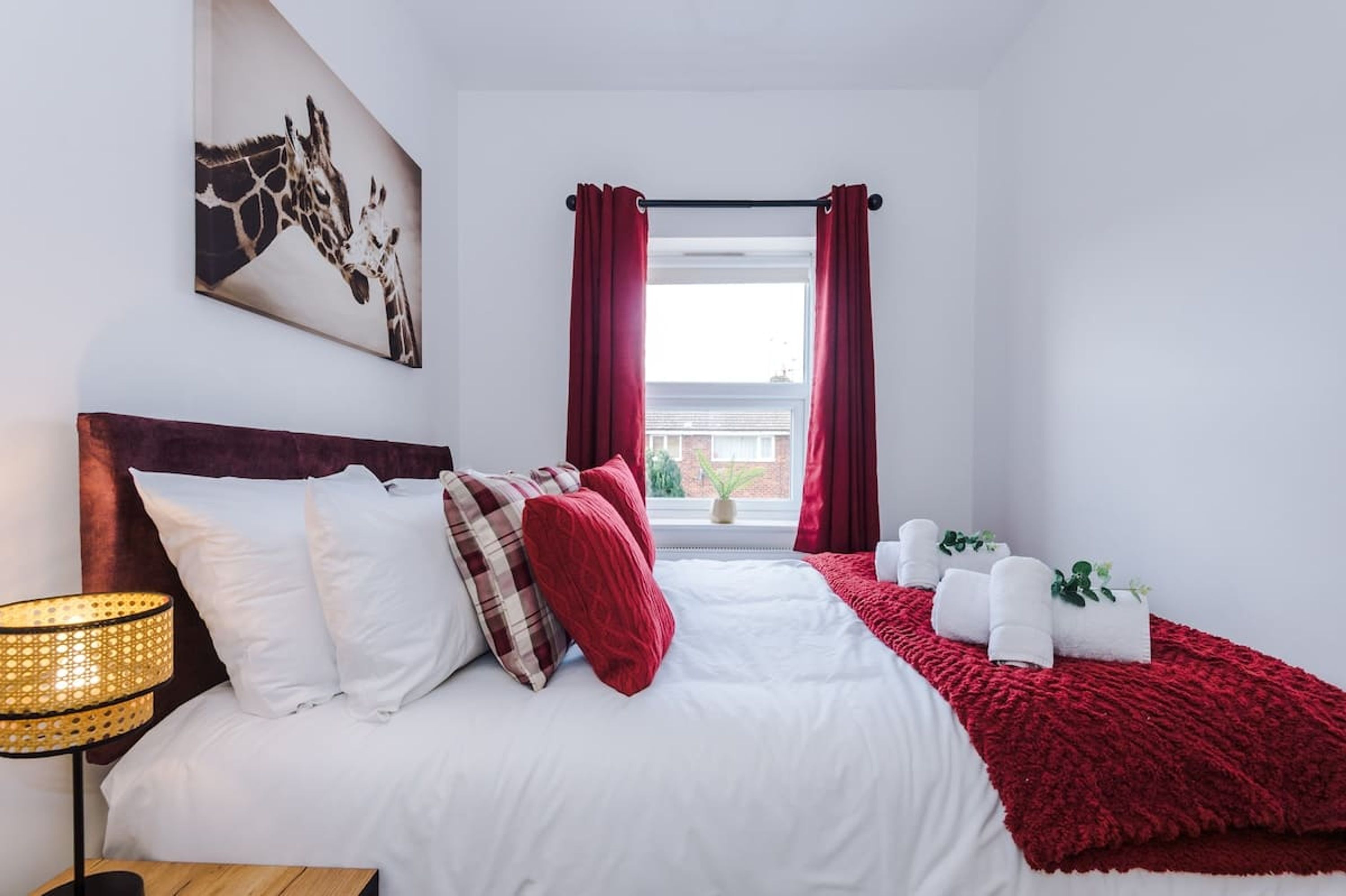 A bedroom with a bed, pillows, a lamp, a window with curtains, and a towel rack with towels.