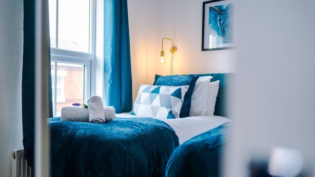 A bedroom with a bed, a nightstand, a wall-mounted lamp, and a window with curtains. - Stunning Home Perfect for Groups Free Parking (Chester)