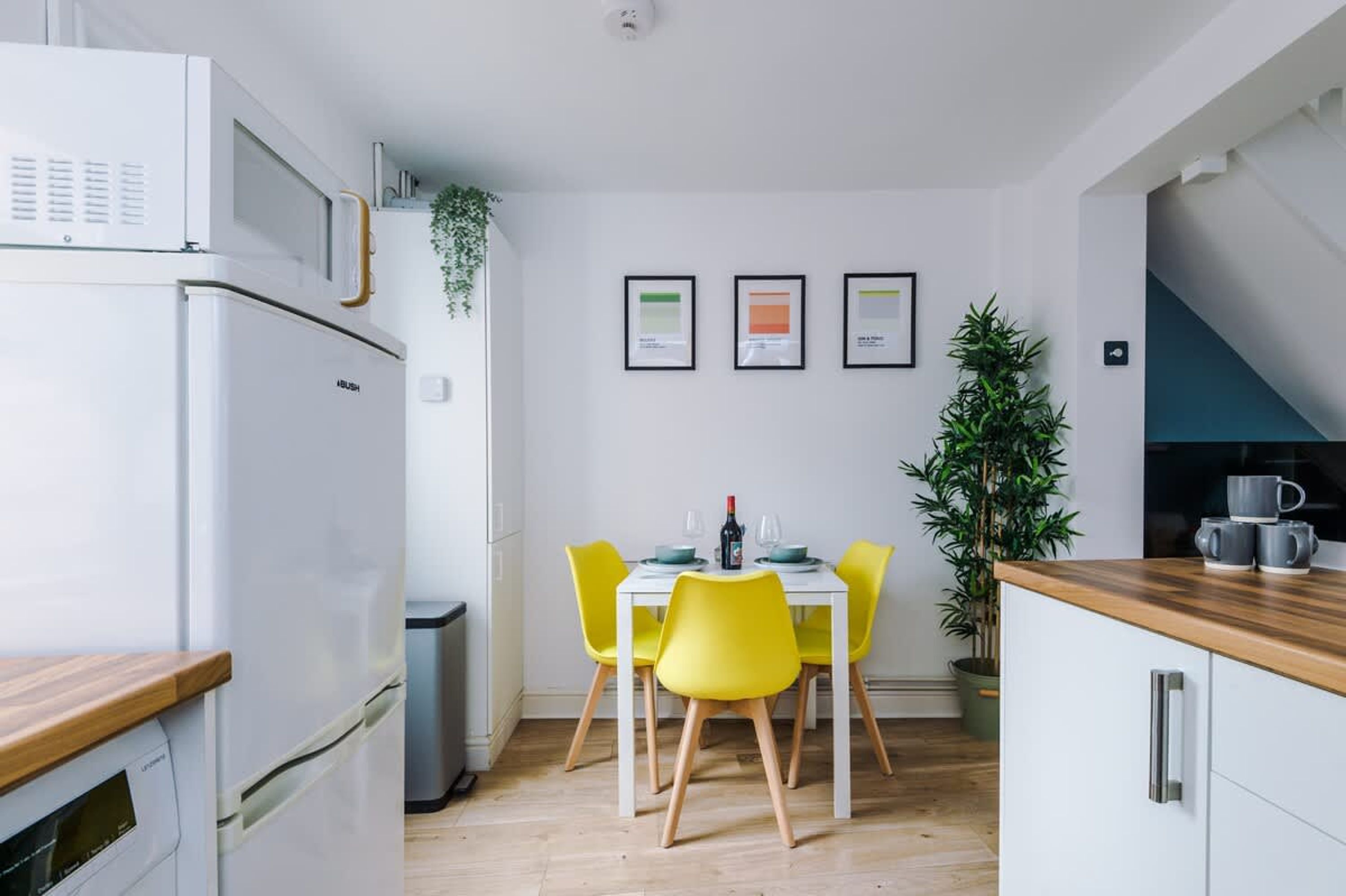 Une cuisine moderne avec un réfrigérateur blanc, des chaises jaunes, une table à manger et des tableaux encadrés accrochés au mur.