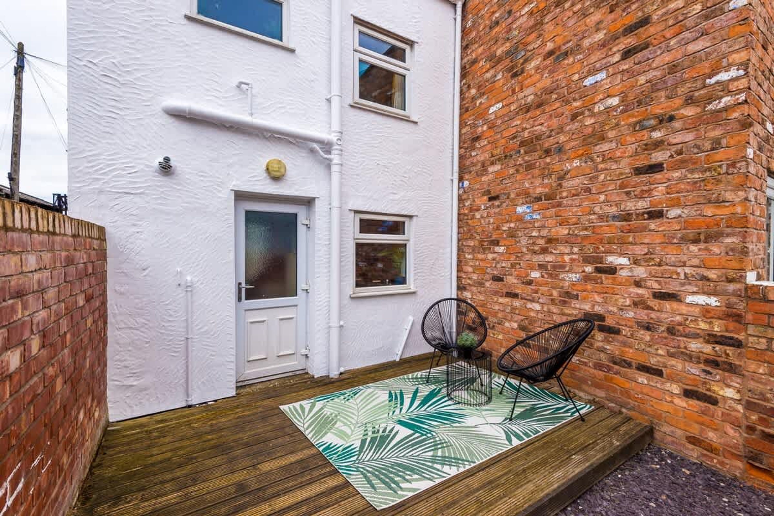 A small courtyard with a white door, a brick wall, and two black chairs on a patterned rug.