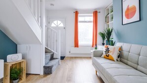 A modern living room with a staircase, a sofa, a radiator, and a window with orange curtains. - Stylish Home Incredible Location Sleeps 5 (Chester)