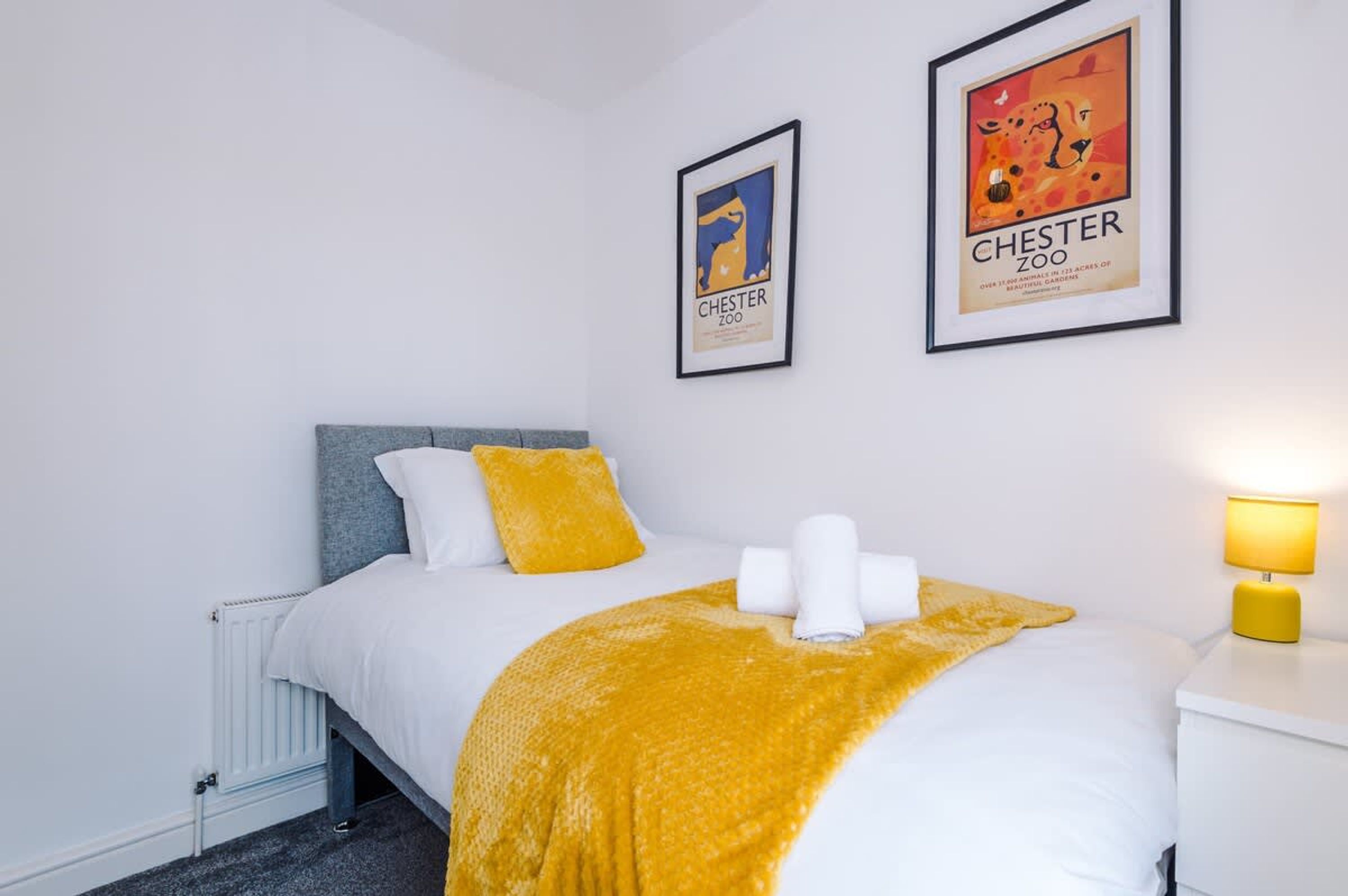 A hotel room with a bed, a radiator, a nightstand with a lamp, and two framed pictures on the wall.