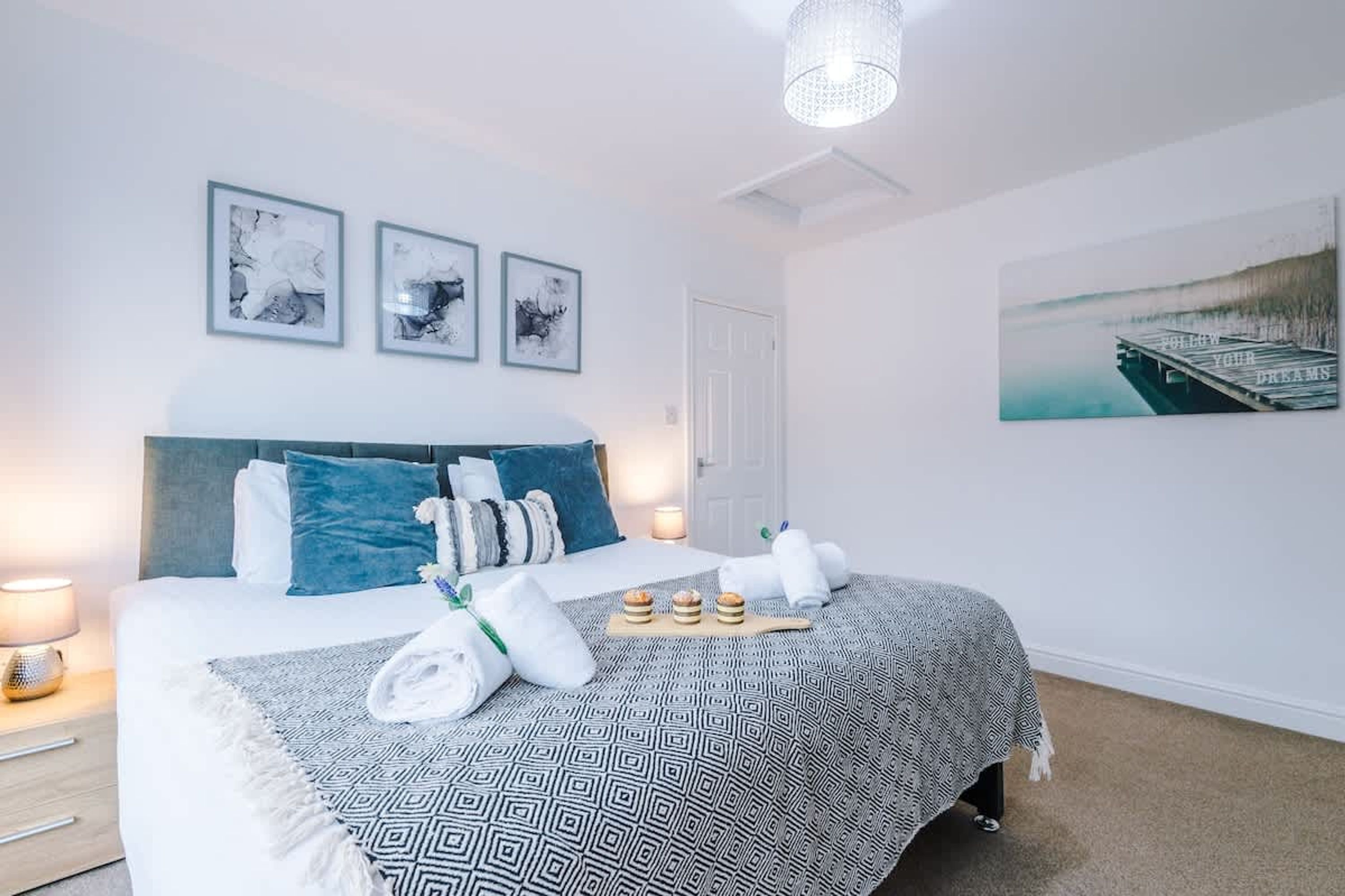 A neatly arranged hotel room with a bed, pillows, a bedside lamp, a framed picture, and a wall-mounted artwork.
