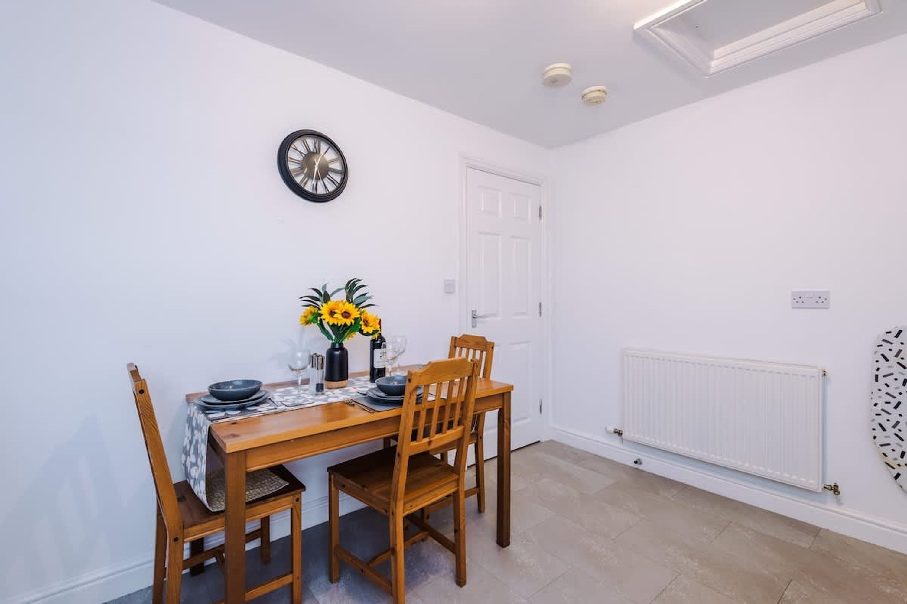 Un coin repas avec une table en bois, deux chaises, une horloge au mur, un radiateur et une porte blanche.