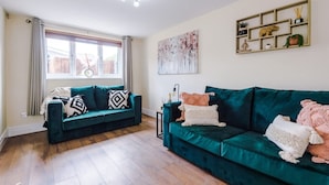 Ein Wohnzimmer mit zwei grünen Sofas, einem fenster mit Vorhängen, einer Wandregal mit Dekoration und einer Deckenleuchte.