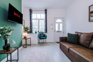 A living room with a brown leather sofa, a teal armchair, a small table with a lamp, a TV mounted on the wall, and a window with curtains. - Stunning 3-bed House Amazing Location Sleeps 5 (Nantwich)