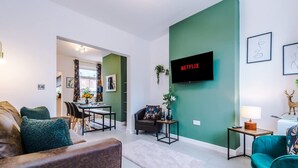 A modern living room with a flat-screen TV, a sofa, a teal armchair, and a dining area with a table and chairs. - Stunning 3-bed House Amazing Location Sleeps 5 (Nantwich)