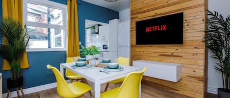 A modern dining area with a white table, yellow chairs, and a wooden wall with a Netflix screen.