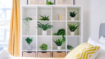 Uma estante branca com vários vasos de plantas, uma moldura com a foto do Chester Zoo e uma cama com almofada amarela e branca.