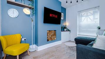 Uma sala de estar com parede azul, uma cadeira amarela, um sofá escuro, uma TV exibindo 'NETFLIX', um relógio, um abajur e uma janela com persianas.