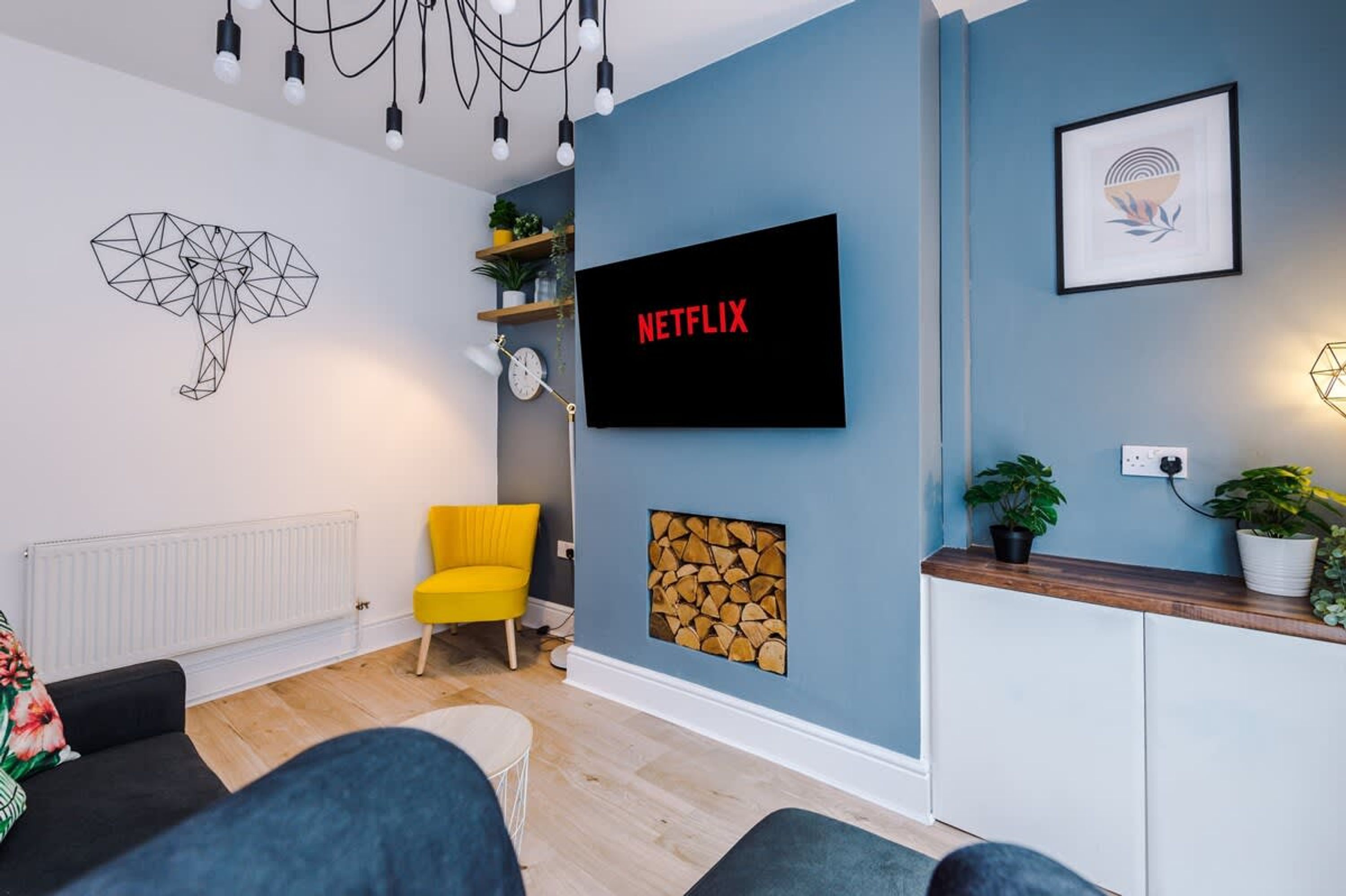 A modern living room with a sofa, a yellow chair, a TV mounted on the wall, a geometric wall art piece, and a wooden cabinet with plants.